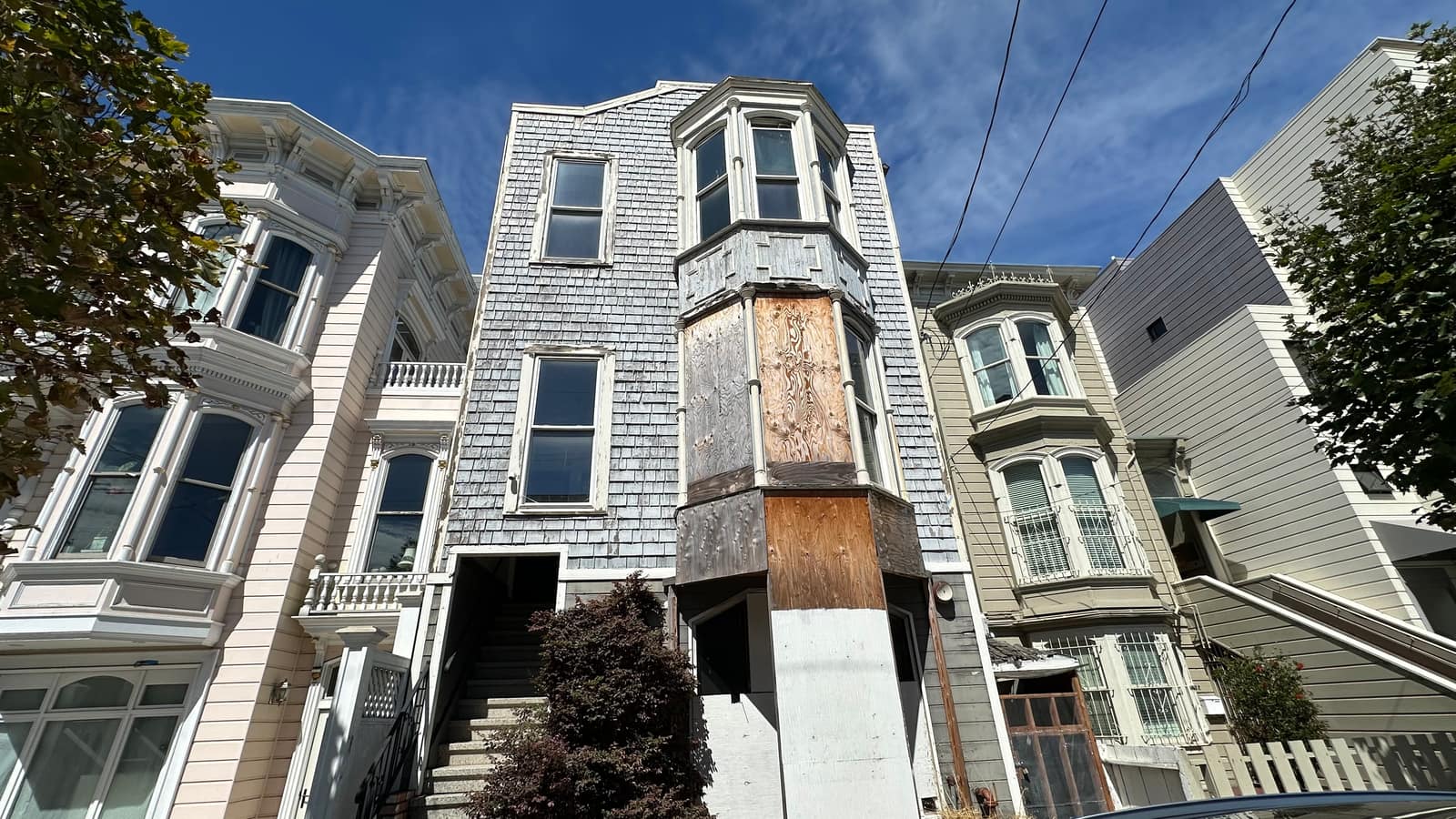 A shingled Victorian with its bay window boarded over in raw plywood, standing between two freshly painted neighbors.