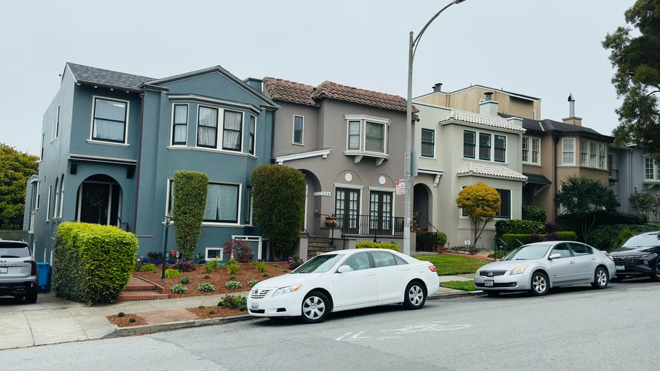 A row of pale blue and grey Edwardian houses along a level street with cars parked outside.