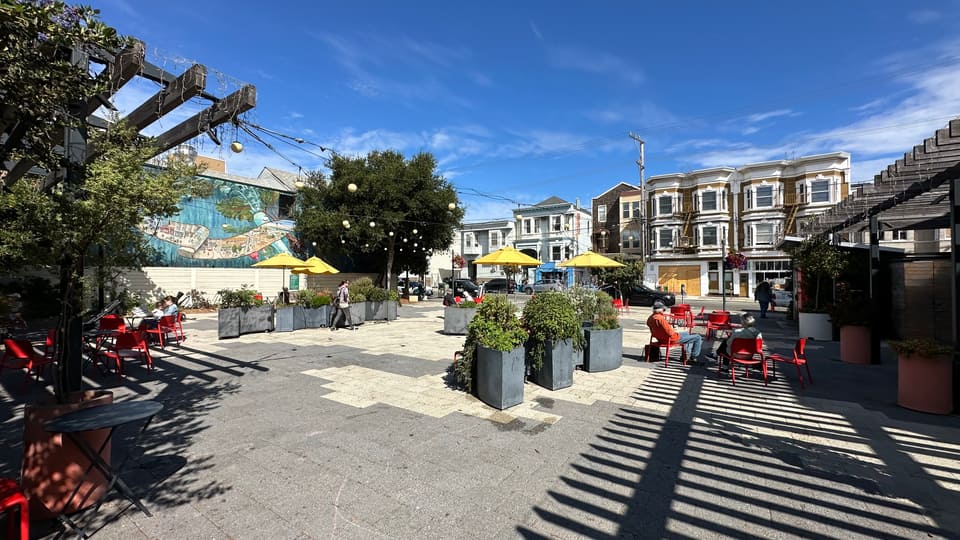 The Noe Valley Town Square seen from the street, pergola and umbrellas above red seating.