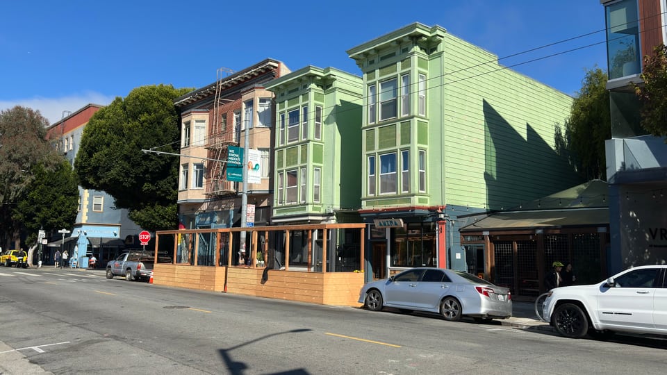 A green Victorian above Hayes Valley storefronts with a timber parklet along the sidewalk.