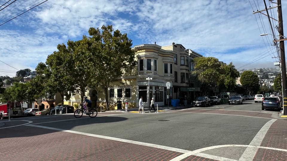 The 24th Street corner in Noe Valley, a Victorian above street-level shops with a cyclist crossing.