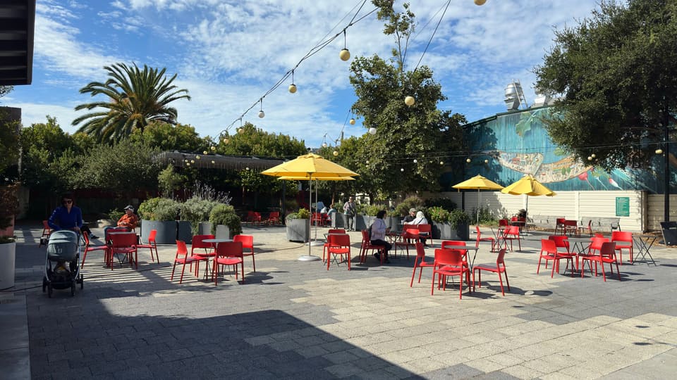 The Noe Valley Town Square parklet, red cafe chairs and yellow umbrellas under a painted mural wall.
