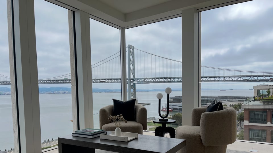A corner-glazed condominium sitting room with boucle armchairs, the Bay Bridge filling the window wall behind.