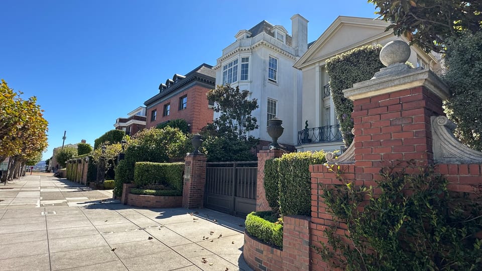A white mansion behind brick gateposts and clipped hedging on an empty tree-lined sidewalk.