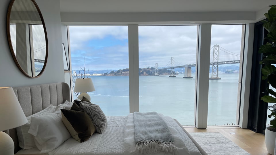 A condominium bedroom with a round mirror and upholstered bed, the Bay Bridge visible through full-height glass.