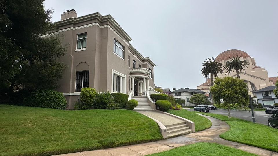 A grey stucco mansion with curved entrance steps and clipped hedging, a domed rooftop rising behind.