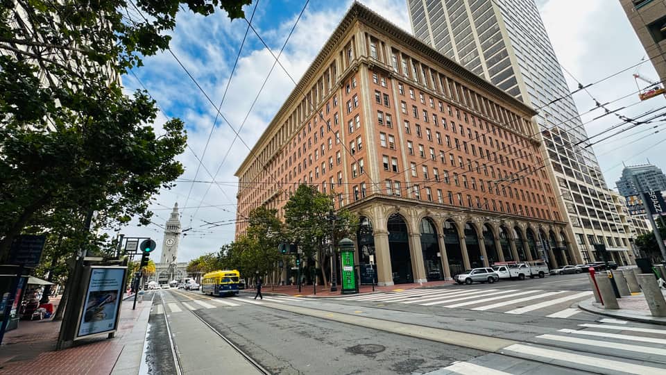 A brick Financial District office block on a wide street, the Ferry Building clock tower in the distance.