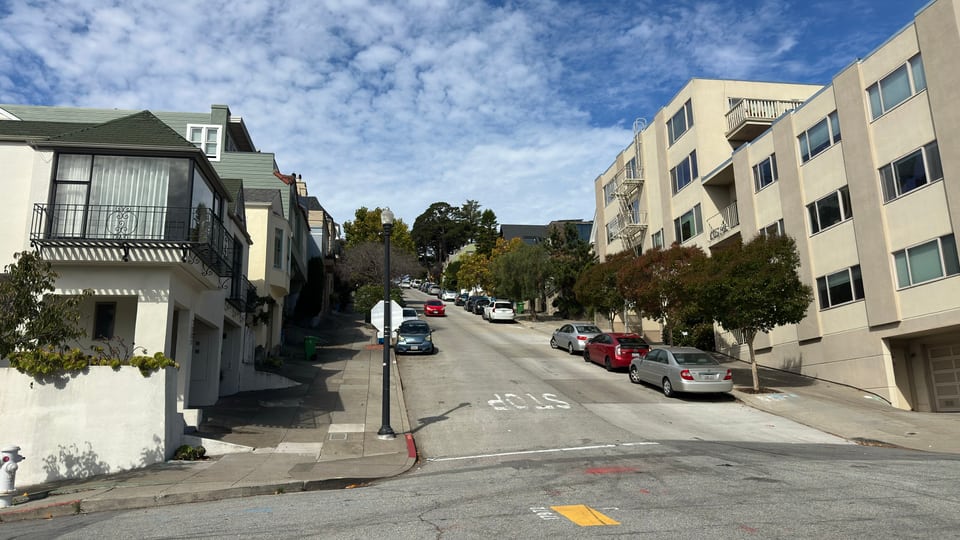 A sloping San Francisco street of stucco apartment buildings with cars parked along both kerbs.