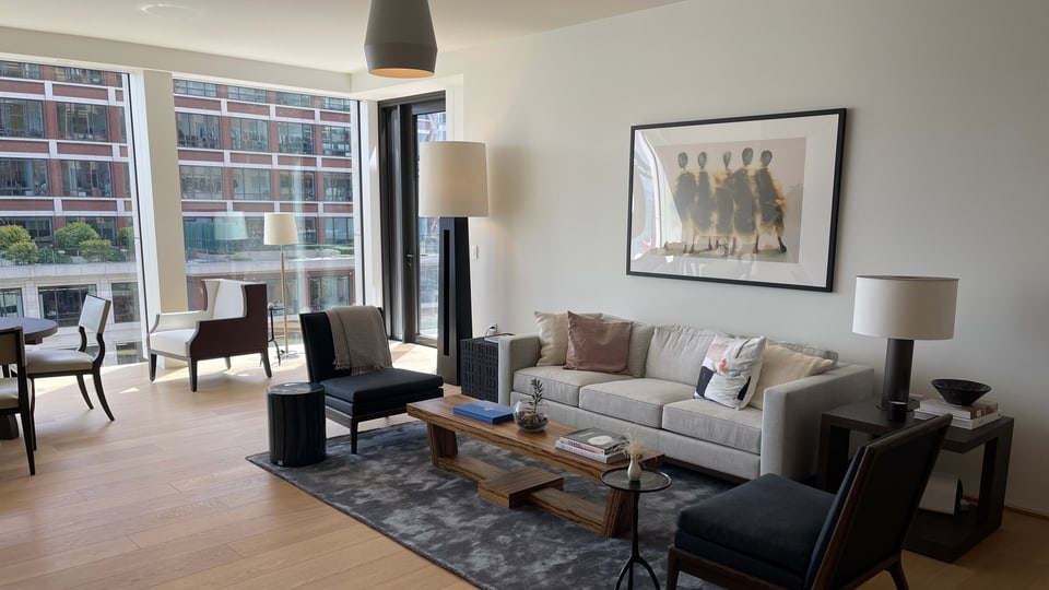 A staged condominium living room with pale sofas, framed artwork and floor-to-ceiling windows onto neighbouring buildings.