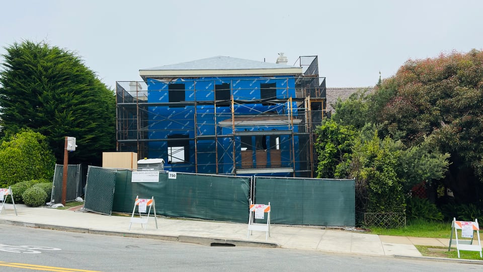 A large house wrapped in blue construction netting and scaffolding behind a green site fence.