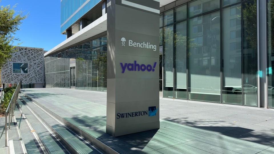 A lobby signage monolith listing technology tenants outside a glass office building, light rail tracks in the foreground.