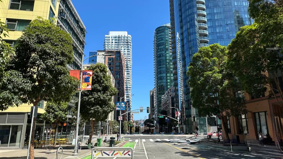 A Transbay street of glass residential towers with young street trees and an empty crosswalk under clear sky.