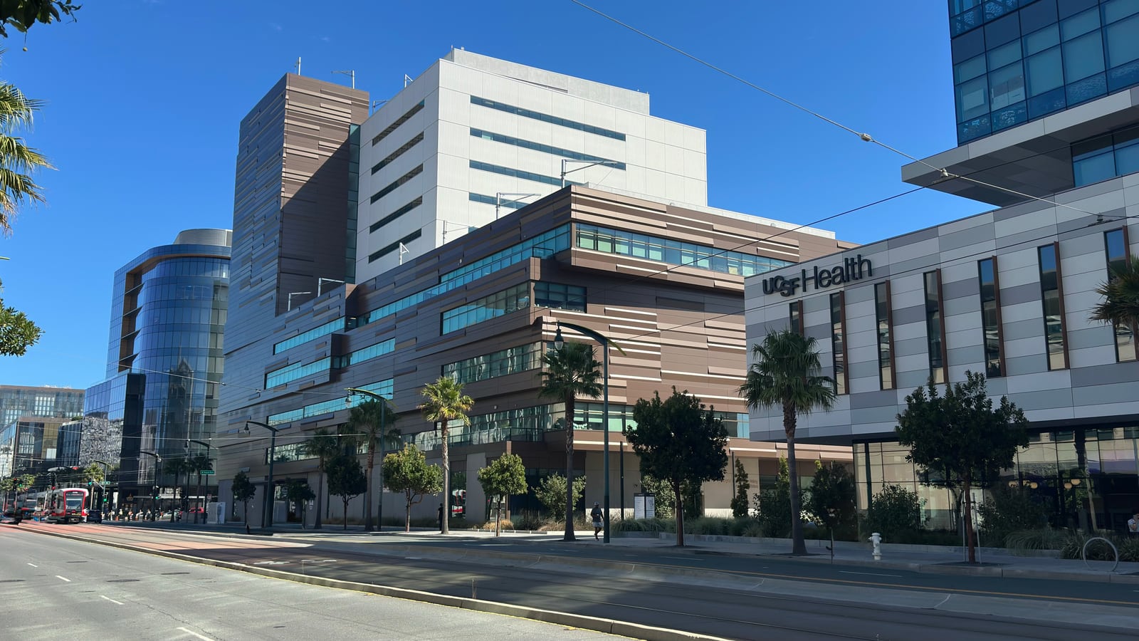 The UCSF Health campus at Mission Bay, its buildings stepping back along Third Street behind palms, a Muni train passing at the left.
