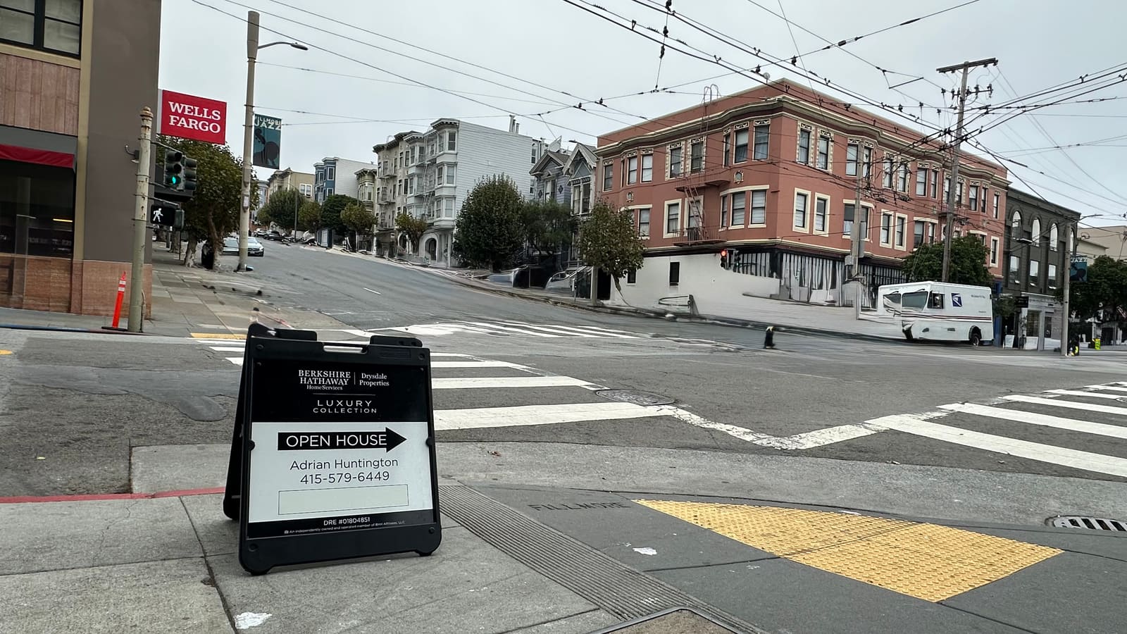 An open-house A-frame sign on a Fillmore Street corner, with Victorian flats climbing the hill behind it on an overcast San Francisco morning.